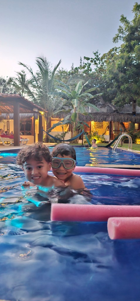 Duas crianças sorrindo na piscina, usando óculos de mergulho, rodeadas por árvores e uma área de descanso ao fundo.