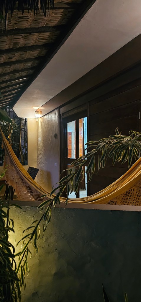 Uma rede amarela pendurada em um ambiente aconchegante, com paredes de madeira e iluminação suave, rodeada por plantas.