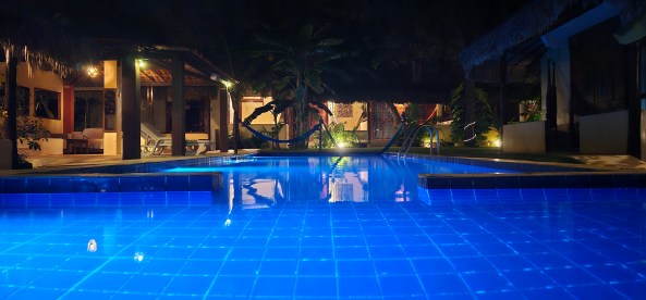 Piscina iluminada à noite, com azulejos azuis, cercada por vegetação e áreas de descanso.