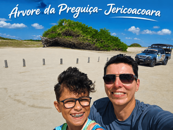 Selfie de duas pessoas sorrindo em frente à Árvore da Preguiça em Jericoacoara, com um céu azul e uma paisagem de areia ao fundo.